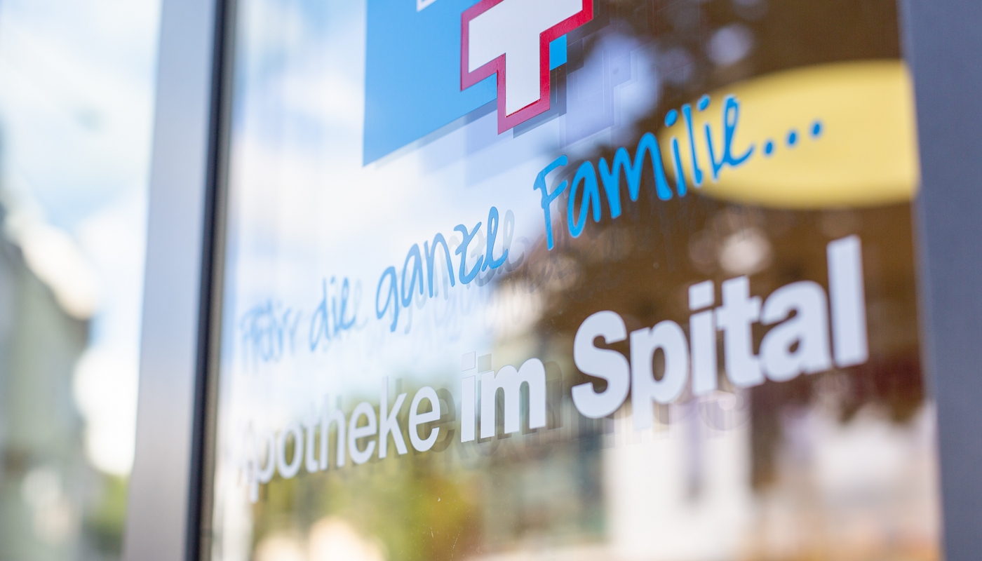 Perspektive auf das Logo der Apotheke im Spital Nahaufnahme des Logos der Apotheke im Spital auf einer Glastür. Der Schriftzug "Für die ganze Familie" hebt die familiäre Atmosphäre und den Service der Apotheke hervor.
