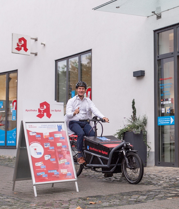 Umweltbewusste Lieferung mit dem Lastenrad Ein Apotheker trägt einen Fahrradhelm und sitzt auf einem Lastenfahrrad vor der Apotheke im Spital. Neben ihm steht ein Werbeaufsteller mit aktuellen Angeboten der Apotheke. Im Hintergrund sind große Fenster und der Eingang der Apotheke zu sehen.