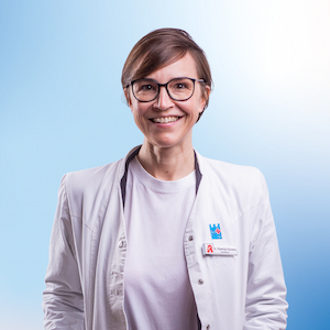 Ksenija Gorenc, eine Apothekerin im weißen Kittel, lächelt herzlich in die Kamera. Sie trägt eine moderne Brille und ein Namensschild mit Apotheken-Logo, welches sie als Mitarbeiterin der Apotheke ausweist. Der Hintergrund ist in einem freundlichen Blauton gehalten, was ein professionelles Erscheinungsbild vermittelt.