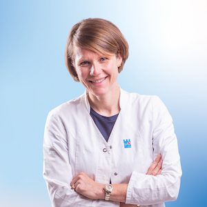 Verena Vetter, eine Apothekerin im weißen Kittel, steht mit verschränkten Armen und einem freundlichen Lächeln vor der Kamera. Sie trägt ein Apotheken-Logo auf ihrem Kittel, das sie als Mitarbeiterin der Apotheke ausweist. Der Hintergrund ist in einem hellen Blauton gestaltet, was ein professionelles und vertrauenswürdiges Erscheinungsbild vermittelt.