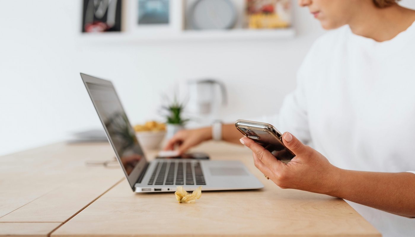 Online Bestellung Eine Frau sitzt an einem Schreibtisch mit einem Laptop und hält ein Smartphone in der Hand. Im Hintergrund sind unscharf Dekorationselemente und Pflanzen sichtbar.