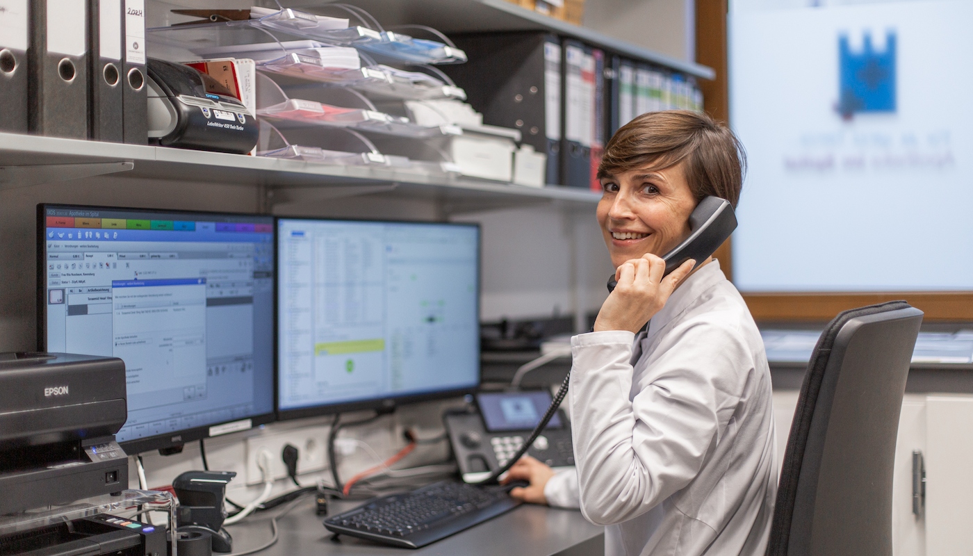 Eine Mitarbeiterin im weißen Kittel sitzt lächelnd in einem Büro der Apotheke und telefoniert.