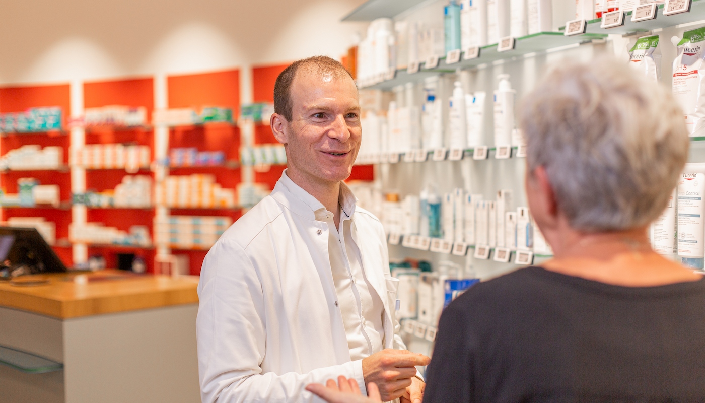Individuelle Beratung durch Ihren Apotheker Ein Apotheker im weißen Kittel berät eine Kundin vor einem Regal mit Pflegeprodukten in einer Apotheke.