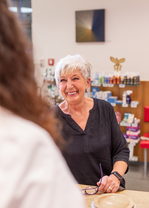 Eine freundlich lächelnde Frau mit weißem Haar steht an einem Apothekentresen und lässt sich beraten.