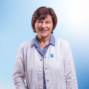 Christine Linde, eine Mitarbeiterin der Apotheke, lächelt herzlich in die Kamera. Sie trägt einen weißen Kittel über einer blauen Bluse mit feinem Muster. Ihr braunes, halblanges Haar ist locker gestylt. Der helle blaue Hintergrund unterstreicht ihre freundliche und professionelle Ausstrahlung.