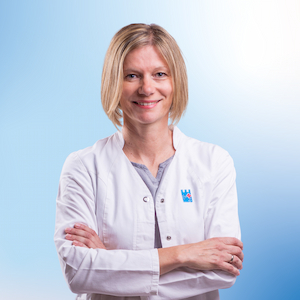 Petra Weiß, eine Mitarbeiterin der Apotheke, lächelt freundlich in die Kamera. Sie trägt einen weißen Kittel mit verschränkten Armen und wirkt professionell und zugänglich. Ihr blondes, kinnlanges Haar ist glatt gestylt. Der blaue Hintergrund gibt dem Bild eine klare und moderne Ausstrahlung.