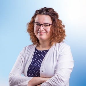 Simone Zodel, eine Mitarbeiterin der Apotheke, trägt einen weißen Kittel und ein dunkelblaues, gepunktetes Oberteil. Sie steht mit verschränkten Armen und einem freundlichen Lächeln vor der Kamera. Der helle, blaue Hintergrund hebt ihr professionelles Erscheinungsbild hervor und verleiht dem Bild eine einladende Atmosphäre.