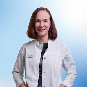 Stephanie Miene, eine Mitarbeiterin der Apotheke, trägt einen weißen Kittel und ein farbenfrohes Oberteil darunter. Sie lächelt freundlich in die Kamera, während ihr schulterlanges, glattes Haar das professionelle Erscheinungsbild unterstreicht. Der blaue Hintergrund verleiht dem Bild eine helle, moderne Ausstrahlung.